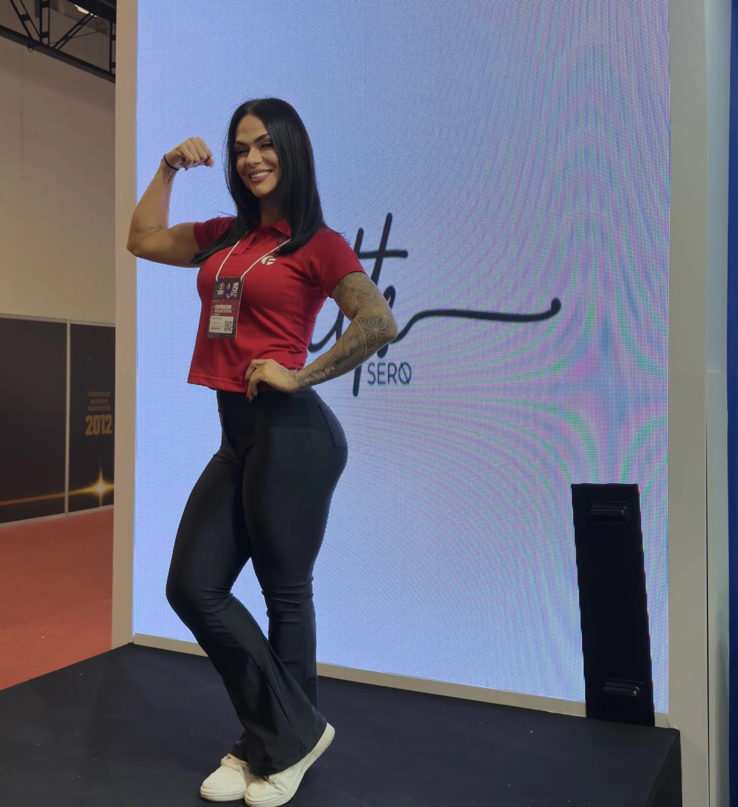 Beatriz Faria em pose de força sobre um palco de feira, de camiseta vermelha