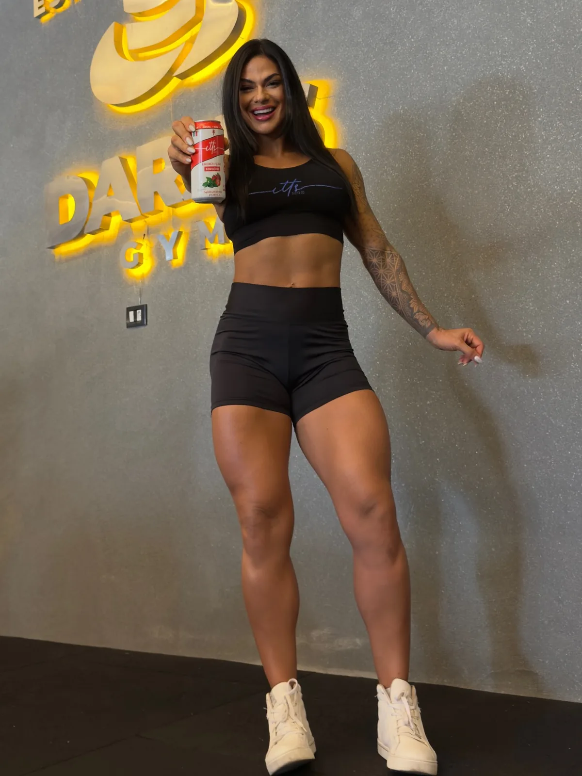 Beatriz Faria em uma academia, de top e shorts pretos, segurando uma lata da ITTS Sero